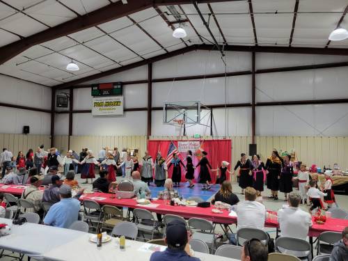 A lively dance performance in a gymnasium, with participants in traditional attire and an audience seated at tables.