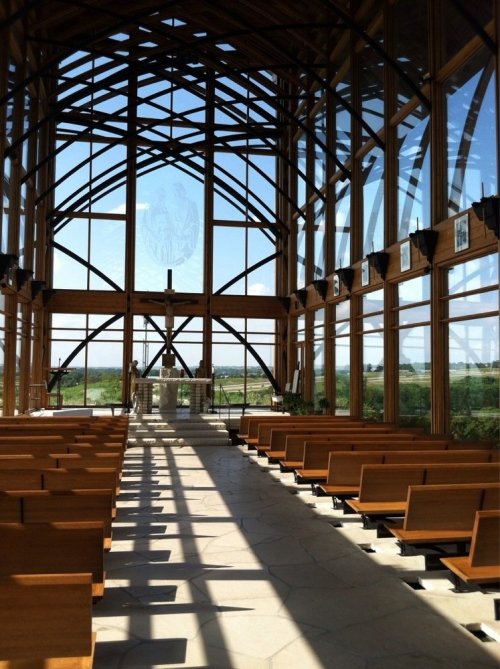 Interior of a modern chapel with wooden beams, large windows, and rows of wooden benches, bathed in natural light.