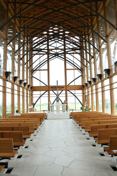 Interior of a modern church with wooden beams, large windows, and rows of wooden benches facing an altar.
