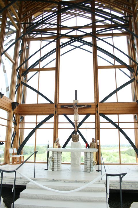 Interior of a modern chapel with wooden beams, a crucifix, and an altar, featuring large windows and a scenic view.