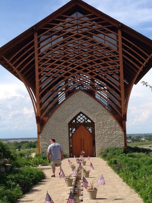 A man walks towards a modern wooden chapel with large glass windows, flanked by potted plants and small American flags.