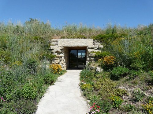 A stone entrance set into a grassy hillside, surrounded by colorful plants and flowers under a clear blue sky.