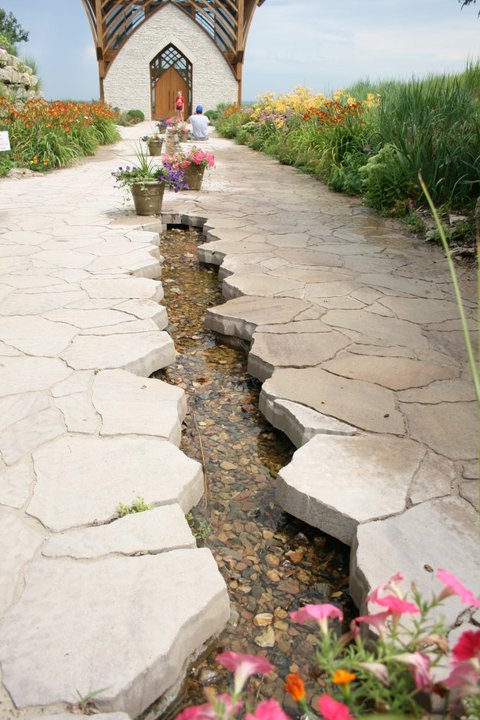 A stone pathway with a water feature, surrounded by colorful flowers and leading to a building in the background.