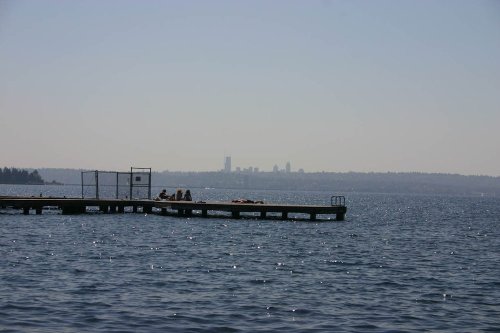 A distant view of a city skyline across a shimmering body of water, with a dock in the foreground and people relaxing.