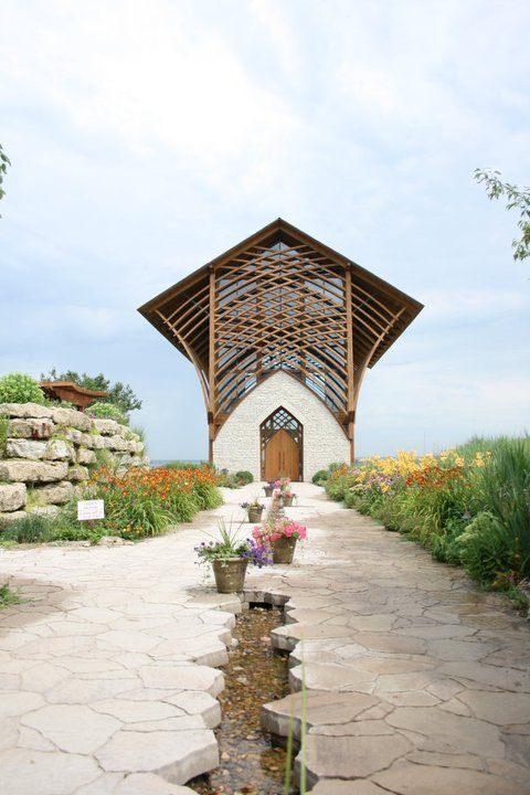 A modern wooden chapel with a large glass front, surrounded by colorful flowers and a stone pathway.