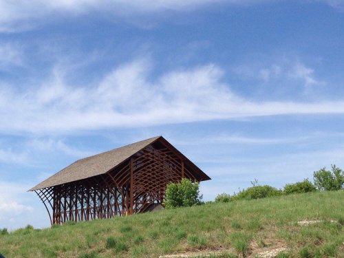 A unique wooden structure with a sloped roof sits on a grassy hill under a blue sky with scattered clouds.