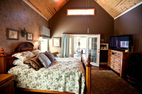 Cozy bedroom with a wooden bed, decorative pillows, and a TV, featuring high ceilings and natural light from a window.