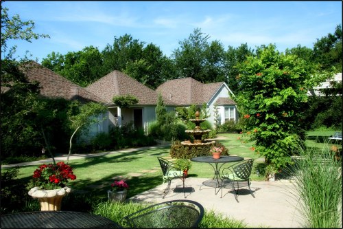 A serene garden scene featuring a fountain, lush greenery, and cozy seating areas surrounded by charming houses.
