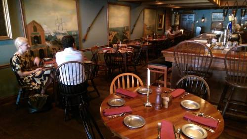 A cozy restaurant interior with wooden tables, chairs, and nautical-themed decor. Two diners are seated in the background.