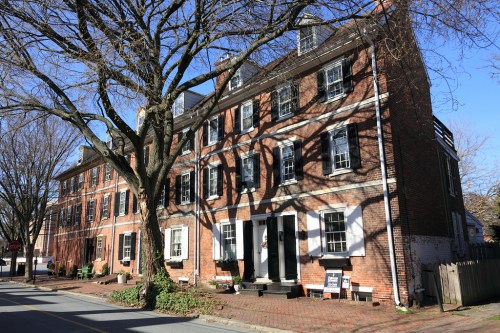 Historic brick building with large windows, surrounded by trees, casting shadows on a sunny day.