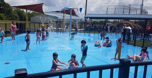 Children playing in a splash pad with water features on a sunny day, surrounded by parents and playground equipment.