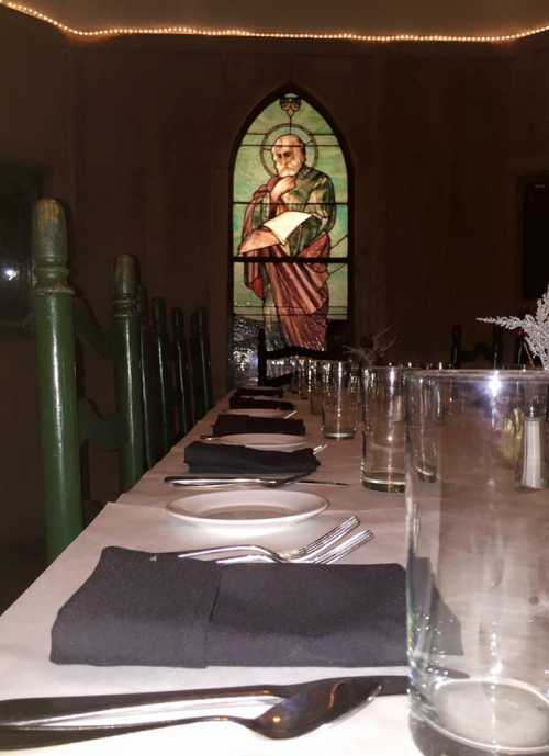 A long dining table set with black napkins and glassware, with a stained glass window of a saint in the background.