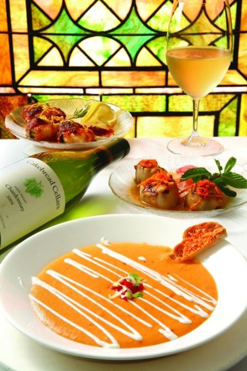 A plate of creamy soup with a drizzle, accompanied by seafood dishes and a glass of white wine on a decorative table.