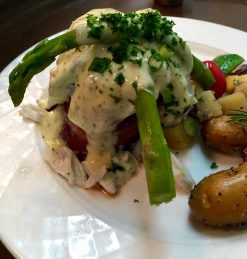 A plate of chicken topped with creamy sauce, asparagus, and herbs, served with roasted potatoes and cherry tomatoes.