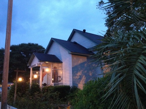 A cozy house at dusk, featuring a porch with lights and an American flag, surrounded by greenery and palm trees.