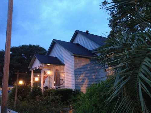 A cozy house at dusk, featuring a porch with lights and an American flag, surrounded by greenery and palm trees.