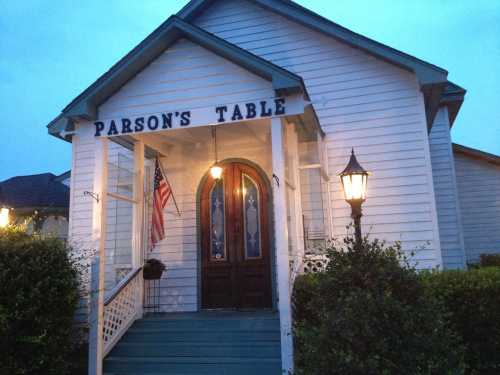 A charming white house with a sign reading "Parson's Table," featuring a porch and a lamp post, under a twilight sky.