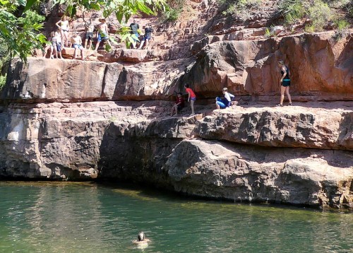 A group of people on rocky cliffs by a green swimming hole, with one person swimming and others exploring the area.
