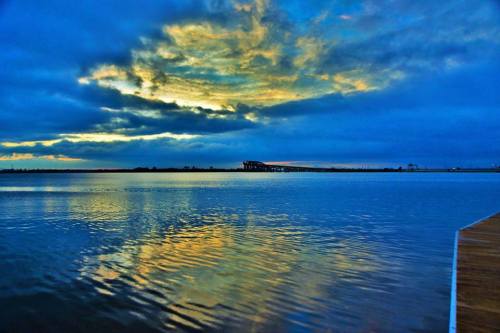 A serene lake at sunset, reflecting vibrant clouds and colors in the water, with a distant structure on the horizon.