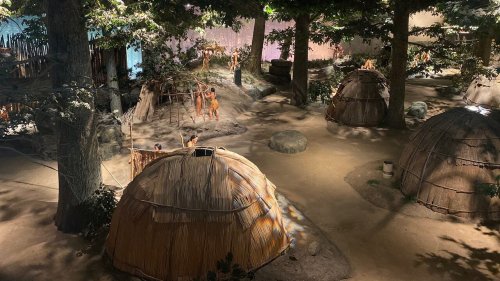 A historical village scene with thatched huts, trees, and figures engaged in activities in a natural setting.