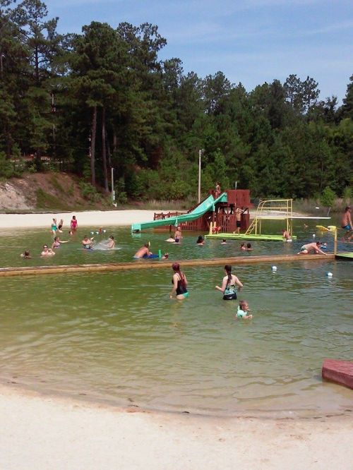 A sandy beach area with a green water slide, people swimming, and children playing in a natural pond surrounded by trees.