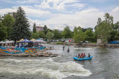 A group of people rafting on a river near a colorful riverside café and lush greenery under a cloudy sky.