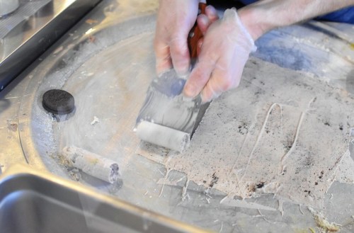 A person in gloves uses a metal spatula to roll ice cream on a cold surface, with some ingredients nearby.