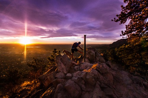 A hiker climbs a rocky outcrop at sunset, with vibrant colors illuminating the sky and landscape below.