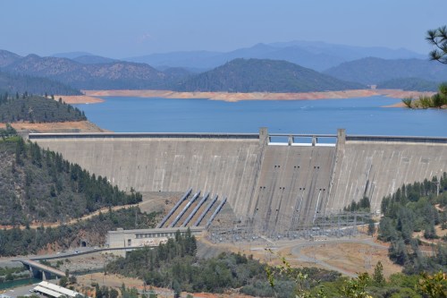 A large concrete dam with spillways, surrounded by trees and mountains, overlooking a blue lake under a clear sky.