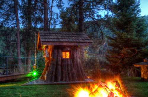 A cozy treehouse with a glowing window, surrounded by trees and a campfire in the foreground.