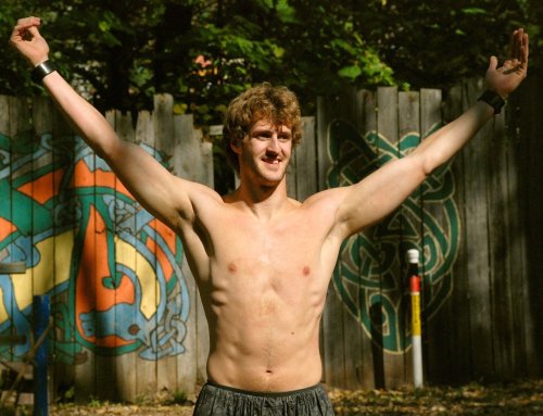 A shirtless man with arms raised stands outdoors in front of a colorful mural on a wooden fence.