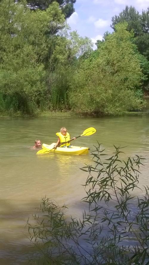 A person in a yellow kayak paddles on a calm river, while another person swims nearby among green trees.