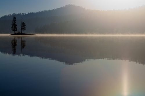 A serene lake at dawn, with mist rising and a small island featuring two trees, surrounded by mountains in the background.