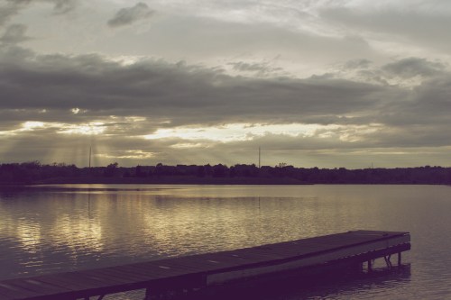 A serene lake at sunset, with a wooden dock extending into the water and clouds reflecting on the surface.
