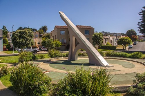A large sundial sculpture surrounded by greenery and residential buildings under a clear blue sky.