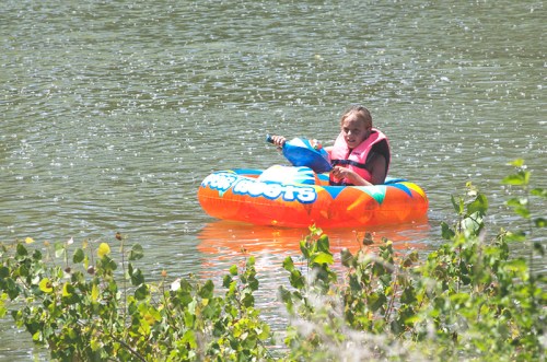 A child floats on an orange inflatable tube in a lake, surrounded by greenery and light rain.