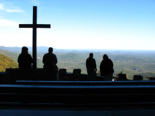 Silhouettes of four people standing by a cross, overlooking a scenic mountain view.