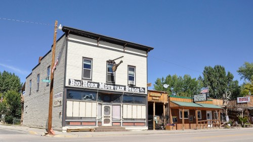 Historic building with a sign for "Big Horn Mountain Stage Co" and nearby wooden structures under a clear blue sky.