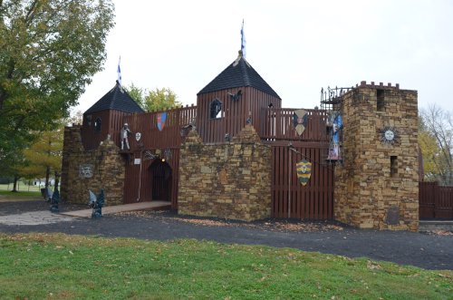 A wooden castle playground with stone walls, towers, and decorative shields, surrounded by trees and fallen leaves.