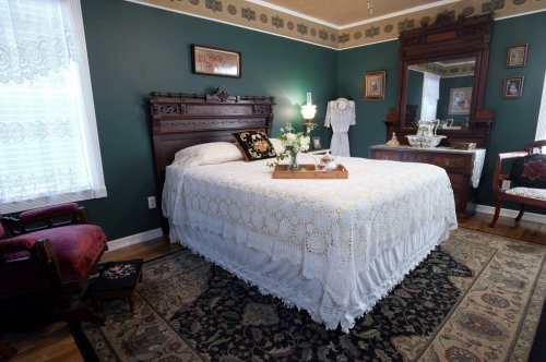A cozy bedroom with a vintage bed, lace bedding, floral decor, and antique furniture against a dark green wall.