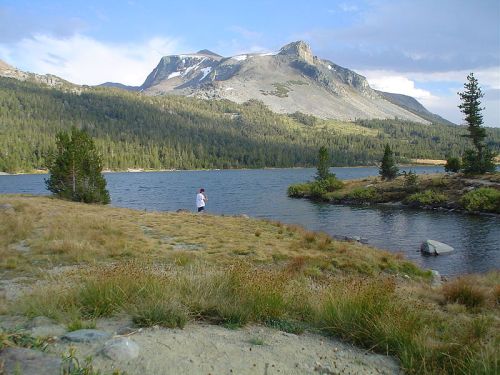 A person stands by a serene lake, surrounded by mountains and lush greenery under a partly cloudy sky.
