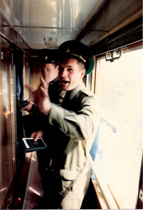 Two uniformed men in a train corridor, one smiling and waving, holding documents. Natural light from windows.