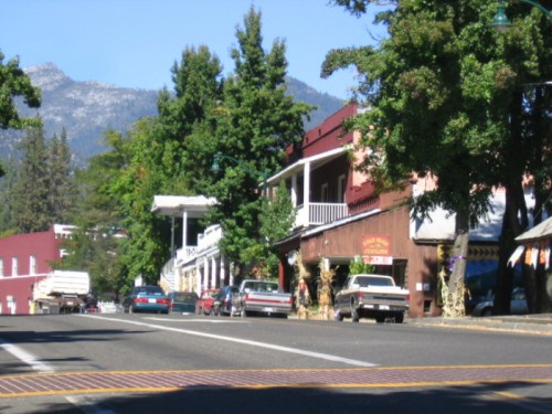 A quiet street lined with trees, shops, and parked cars, with mountains in the background under a clear blue sky.