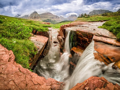 A scenic landscape featuring a waterfall cascading over red rock formations, surrounded by green grass and mountains in the background.