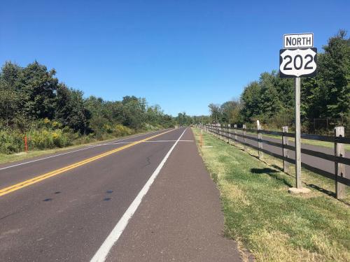 A clear road stretches ahead with a "North 202" sign and a wooden fence alongside green trees and blue sky.