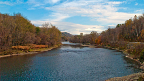 A serene river flows through a landscape of trees with autumn foliage under a blue sky with scattered clouds.