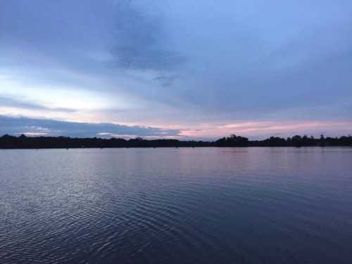 A serene lake at dusk, reflecting soft hues of blue and pink in the sky, surrounded by trees.