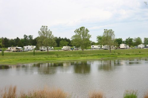 A row of RVs parked near a calm lake, surrounded by green trees and grass under a cloudy sky.