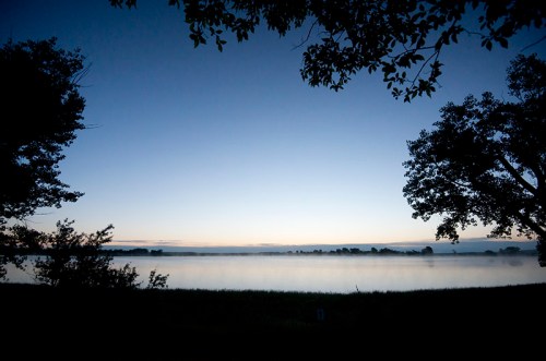 A serene dawn landscape with mist over a calm lake, framed by silhouettes of trees against a blue sky.