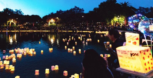 People gather by a lake at dusk, releasing glowing lanterns into the water, creating a serene and festive atmosphere.